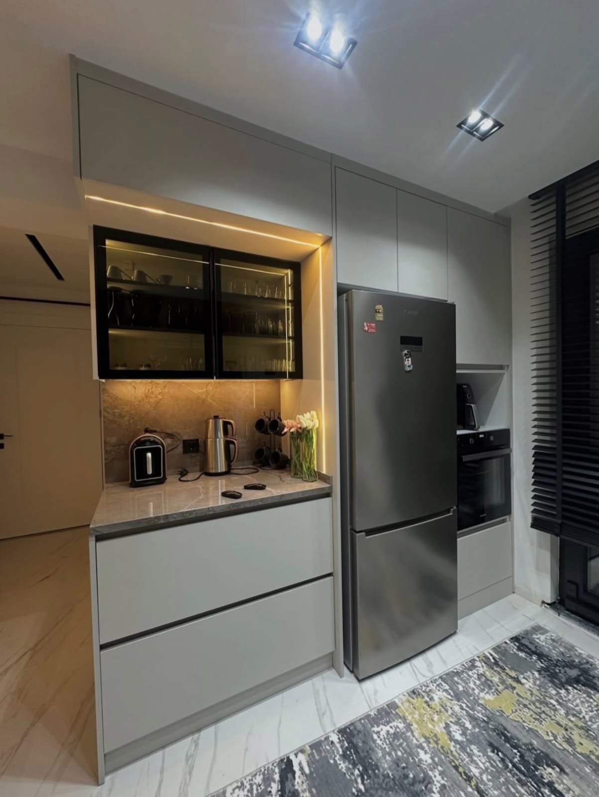 Warm grey cabinetry, glass-front display cabinet with internal lighting, marble-look countertops, and integrated appliances. Practical yet premium. 🍳